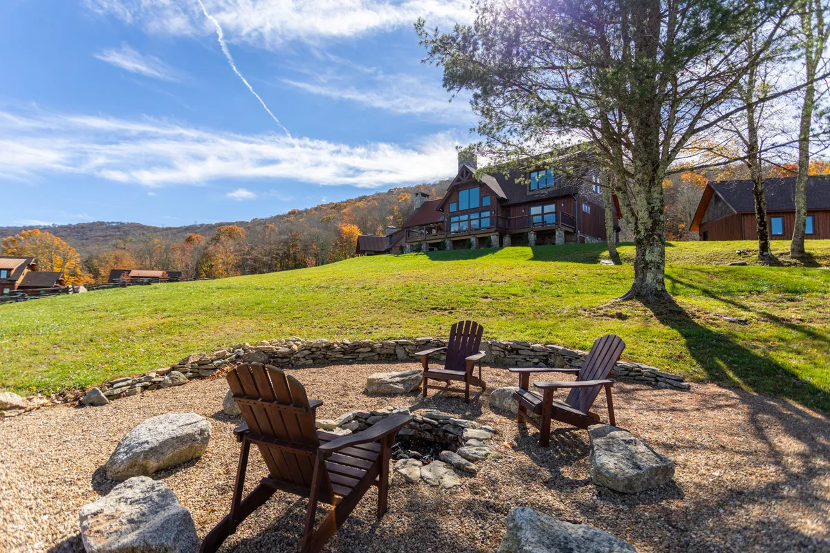 An outdoor scene centered around a stone fire pit encircled by Adirondack chairs, with sweeping views of the Blue Ridge mountains and the main house in the background. The setting is cozy and ideal for evening gatherings under the sky, with the mountains enhancing the atmosphere.