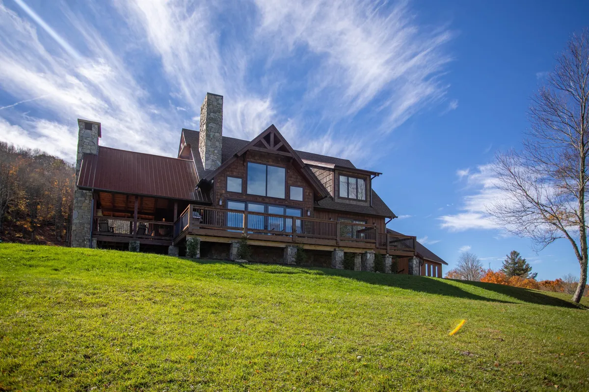 The image features the striking exterior of a modern mountain home, showcasing expansive windows that frame sweeping mountain views, a stately stone chimney, and a sprawling deck set up for gatherings. The scene is bathed in warm sunset light, inviting guests to relax outdoors and enjoy the natural beauty of Banner Elk.