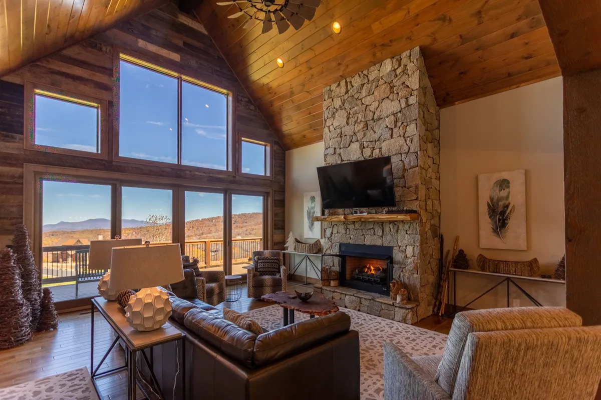 A cozy living room scene centered around a grand stone fireplace, with tall windows that frame mountain vistas. Comfortable seating is arranged to encourage conversation or relaxation by the fire, and natural light fills the space.