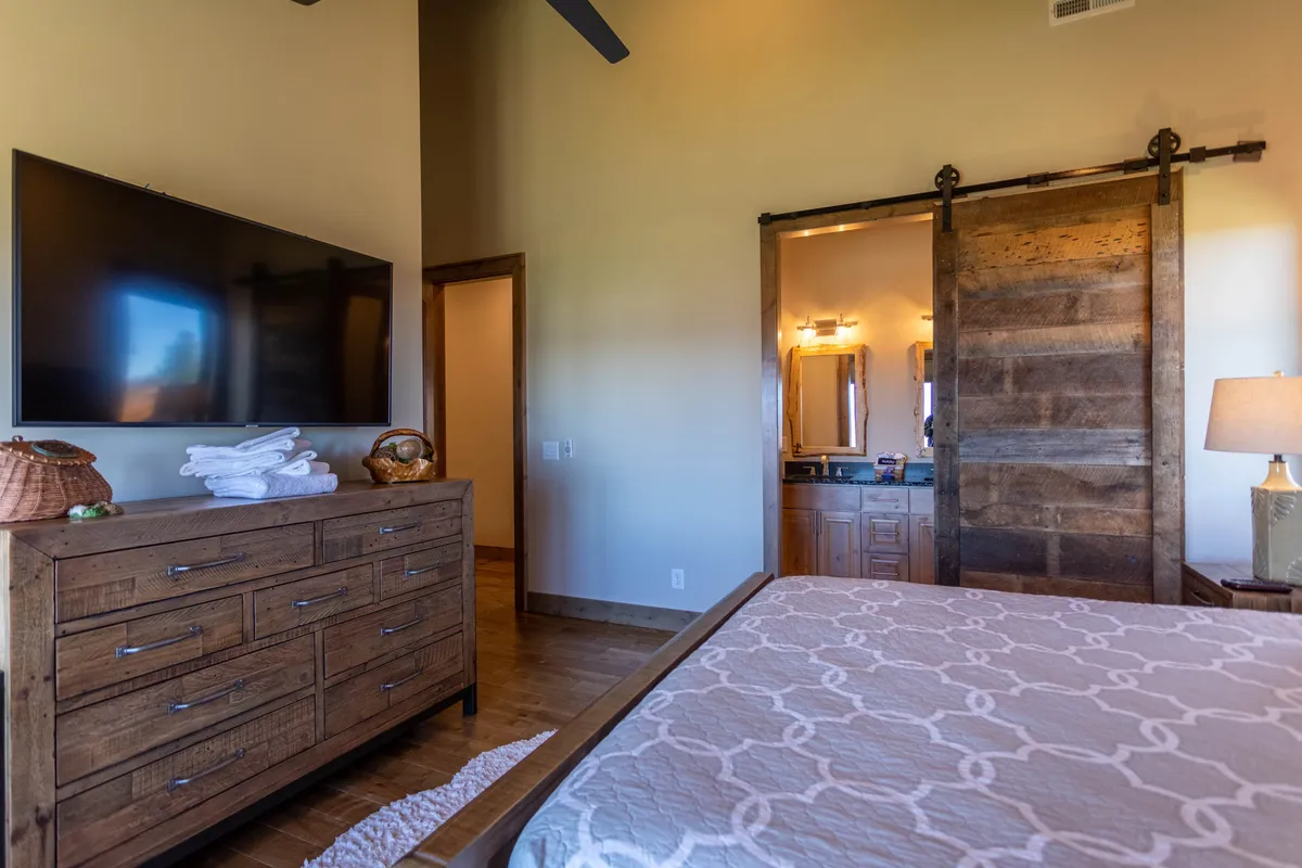 A cozy bedroom scene with a large television, wooden dresser, and an inviting barn door leading to a bathroom. The soft natural light and comfortable furnishings make the space perfect for relaxation or winding down after a long day.