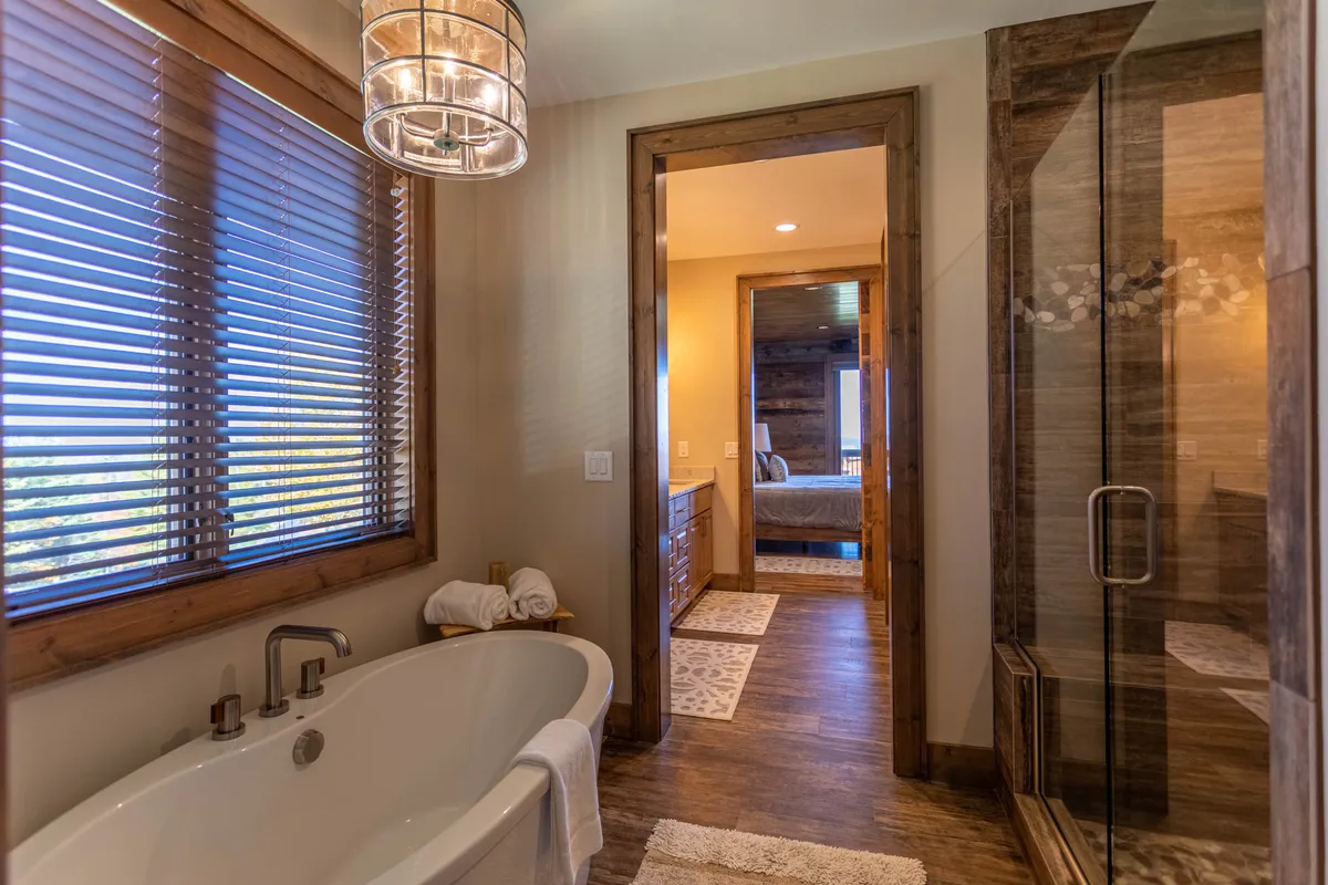 A spacious, spa-like bathroom with a freestanding soaking tub, walk-in shower, wooden vanity, and a large window. The soft light and elegant finishes evoke a sense of relaxation and high-end comfort.