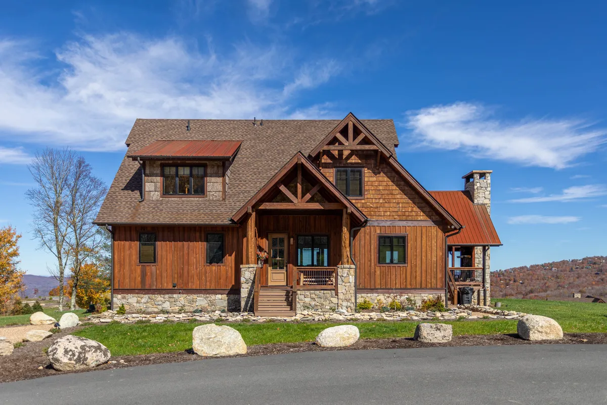 A welcoming exterior view of the property featuring a spacious wooden front porch, stone foundation, dormer windows, and a prominent chimney. The setting captures the home nestled in nature, with gathering spaces perfect for social interaction and relaxation.