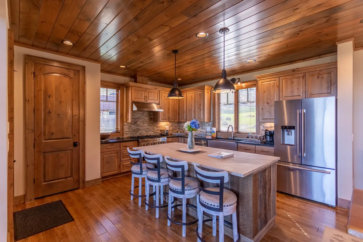 A close-up of the kitchen area emphasizing the large island with four stools, stainless steel appliances, and warm wooden cabinetry. Pendant lights hang above the island, and the layout invites conversation and culinary creativity.