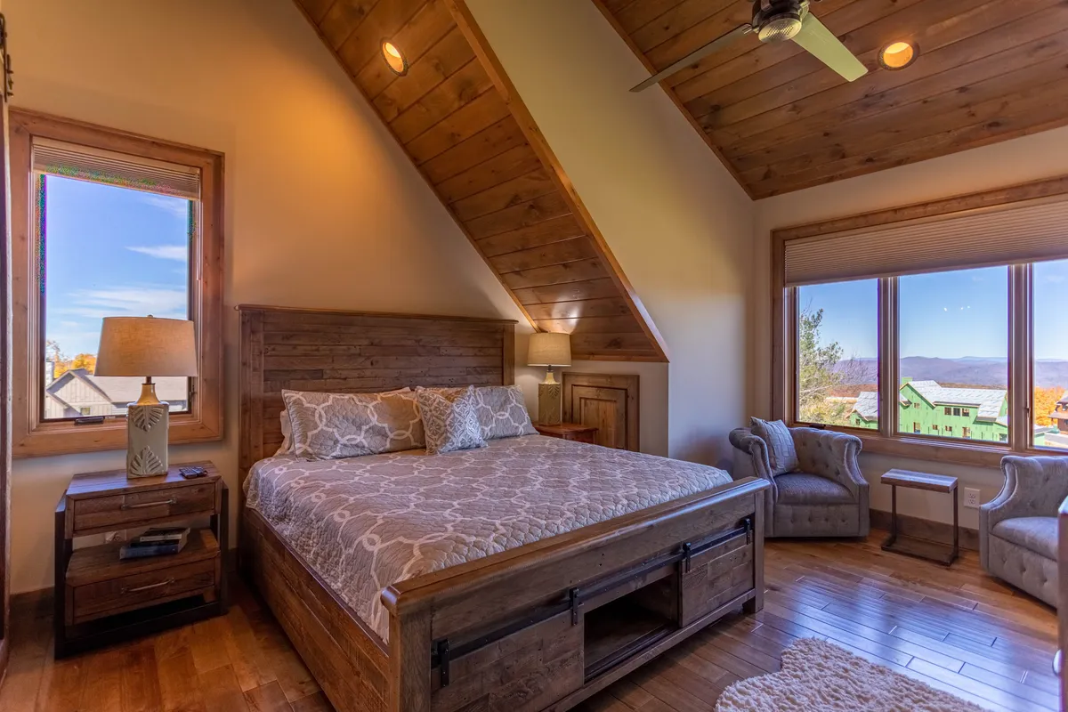 A cozy bed is situated next to a large window with a scenic view, surrounded by warm wooden accents and a softly glowing lamp. The window frames the outdoors, offering a tranquil vantage point for observing the natural environment in any season.