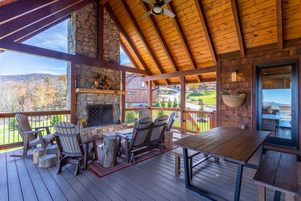 This image features an inviting outdoor living space with a substantial stone fireplace set under a wood-beamed covered deck. There is comfortable cushioned seating arranged for conversation, and a dining table for al fresco meals. The area is designed for relaxation and entertaining, while framing beautiful mountain views in the distance.
