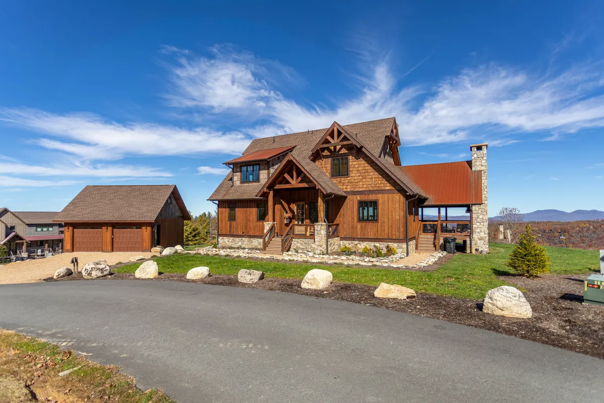 A broad view of the main house, stone chimney, garage, and a lush landscaped garden. The porch and fire pit area invite family gatherings, outdoor meals, and evenings under the stars.