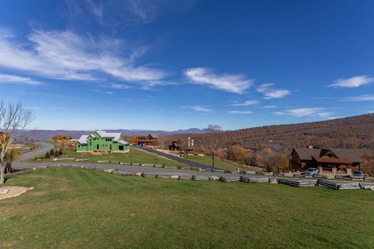 This exterior image captures a green house set against a stunning mountain range, with a winding road, surrounding trees, and neighboring cabins visible. The serene natural beauty, open space, and sense of adventure are prominent, echoing the community’s proximity to mountain trails and group activities.