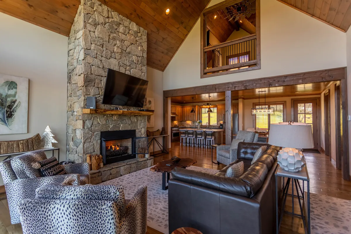 A warm living room setting featuring a stone fireplace, large TV, modern comfortable furniture, and exposed wooden beams. The space is designed for relaxing by the fire, watching movies, and enjoying time with family or guests.