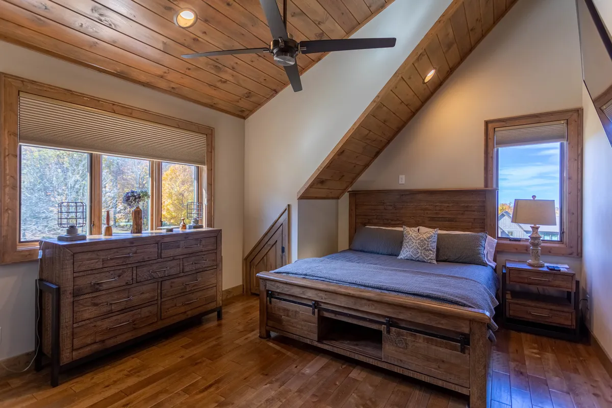 This image features a wooden bed and dresser set in a calm, serene bedroom with large windows that offer clear views outside. The natural light streaming in highlights the tranquil atmosphere and hints at the scenery just beyond the glass, which could include seasonal foliage or snowy landscapes depending on the time of year.