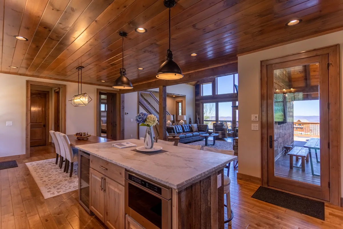 A wide shot capturing the open-concept kitchen, dining area, and living space. A large kitchen island with seating for four is featured in the foreground, with a dining table set for six in the mid-ground. The living area, with a plush sofa and large windows offering a mountain view, is visible in the background. Warm lighting and inviting decor create a welcoming atmosphere.